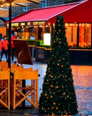 Et juletræ med LED-lys placeret udendørs ved en café eller restaurant. Træet er dekoreret med små varme hvide lys og står ved siden af gul-orange stole under et stort parasol. Omgivelserne har en hyggelig og festlig atmosfære med flere bygninger i baggrunden oplyst med julelys. Træet bidrager til en varm og indbydende stemning på det brostensbelagte område.