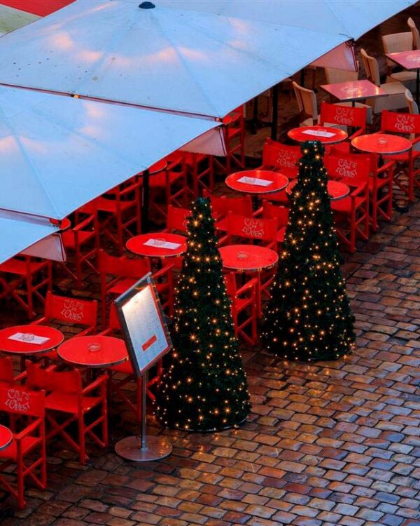 To juletræer med små varme hvide LED-lys står ved siden af røde caféborde og stole under en stor parasol. Træerne er en del af en udendørs juledekoration og er placeret på en brostensbelagt gade. Caféens røde stole og borde skaber en festlig kontrast til det mørke grønne juletræ og de gyldne lys, hvilket giver området en hyggelig og indbydende atmosfære, perfekt til juletiden.