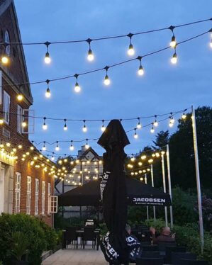 Stemningsfuld udendørs gårdhave med opsatte festoon-lyskæder, der lyser op over borde og parasoller foran historisk bygning under aftenhimmel.