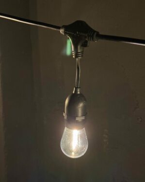 En varm LED-pære hænger fra sort kabel i en T-samling. Lyset spreder sig blødt ud i det mørke rum og skaber hyggelig belysning.