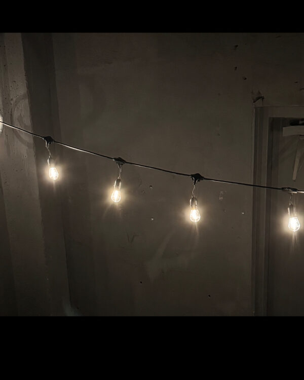 Fem LED-pærer i klar plast hænger fra sort party festoon langs en betonvæg. Lyset er varmt og blødt, og skaber en dæmpet atmosfære.