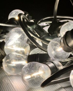 Nærbillede af klare party festoon LED-pærer med sort fatning og kabel, liggende i en bunke. Det varme lys reflekteres i de let ridsede pæreglas.