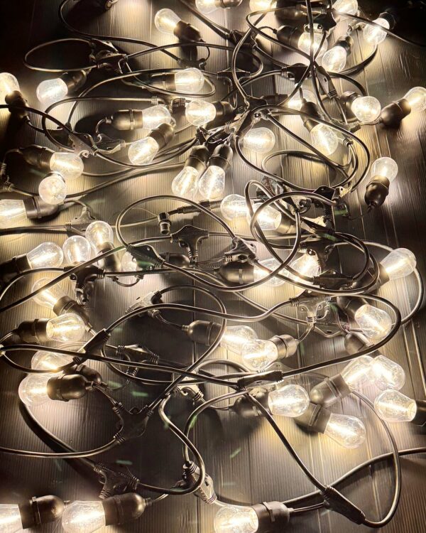 Sammenfiltret net af tændte LED festoon-lyskæder med klare pærer og sorte fatninger, der ligger på sort, riflet underlag og lyser i varm hvid nuance.