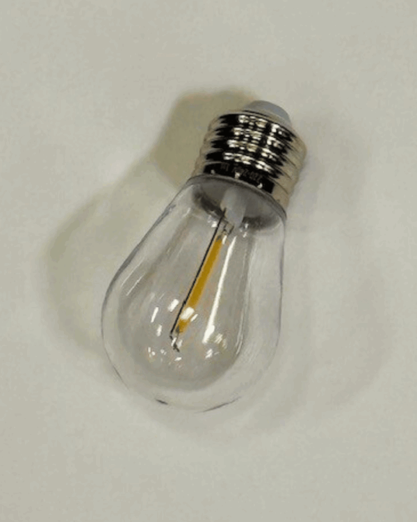 En varm hvid LED-pære med klar glas og E27 fatning, liggende på hvid overflade. Velegnet til udendørs festoon-lyskæder og energieffektiv stemningsbelysning i professionel kvalitet.