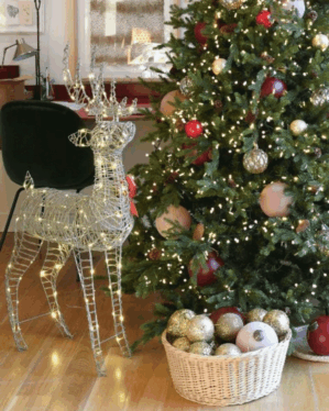 Juletræ med pynt og lys, oplyst rensdyr og kurv med julekugler i hyggelig indendørs scene.