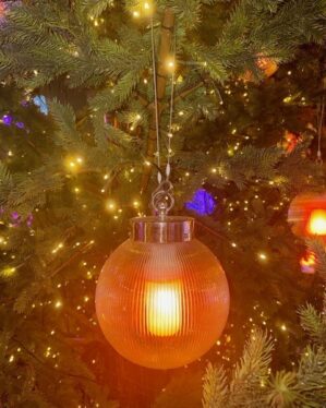 Rund glaslanterne med varm glødende LED-lyskilde hænger i juletræ omgivet af små gule lyskæder, skaber stemningsfuld og elegant belysning til professionel juleudsmykning.