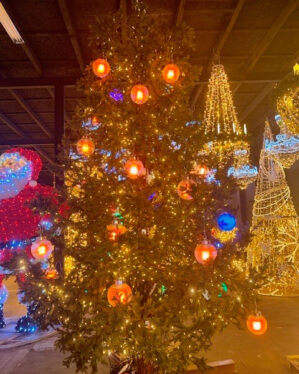 Stort juletræ pyntet med glaslanterner i orange og blå samt små gule LED-lyskæder, udstillet indendørs blandt andre dekorative julemotiver som nisser og lysekroner.
