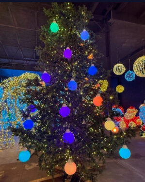 Stort juletræ dekoreret med flerfarvede LED-julekugler i blå, lilla, grøn, gul og rød, kombineret med små varme lyskæder, udstillet indendørs blandt andre lysdekorationer.