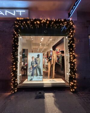 Butiksfacade dekoreret med mørkegrøn gran og varm hvid LED-belysning omkring vinduet. Skaber en elegant og indbydende julestemning foran GANT-butikken i aftentimerne.
