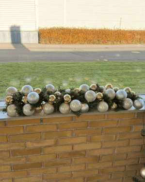 Elegant juleguirlande med guld og sølv julekugler placeret på murstensgelænder. Grønne grene og LED-lys skaber et harmonisk og festligt udtryk i dagslys.