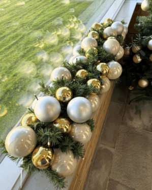 Smuk juleguirlande med guld og sølv julekugler på grønne grene. Placeret på vindueskant med LED-lys, der skaber refleksioner og varm julestemning.