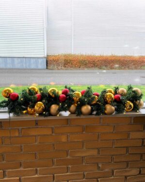 Dekorativ granguirlande pyntet med røde, guld og champagnefarvede julekugler samt små LED-lys. Placeret på murstensvæg udendørs og skaber klassisk juleatmosfære.