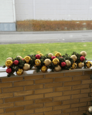 Indendørs granguirlande pyntet med røde, guld og matte julekugler samt små LED-lys. Placeret på murstensvæg og skaber klassisk, stemningsfuld juledekoration.