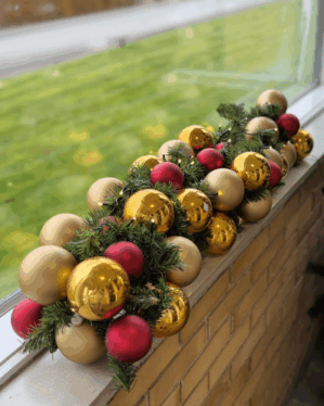 Kunstig granguirlande med røde, gyldne og matte julekugler. Opsat på vindueskant udendørs og pyntet med små LED-lys for klassisk juleudtryk.