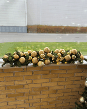 Elegant granguirlande pyntet med matte og skinnende guld julekugler samt små LED-lys. Placeret udendørs på murstensvæg og skaber eksklusiv juleudsmykning.