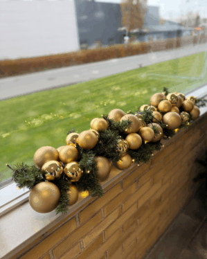 Udendørs juleguirlande pyntet med matte og blanke guld julekugler samt små LED-lys. Placeret på vindueskant og skaber elegant, eksklusiv juledekoration.