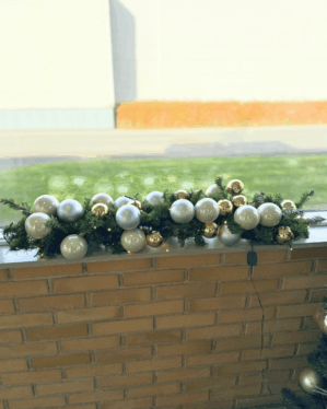 Udendørs juleguirlande pyntet med hvide og gyldne julekugler samt små varme LED-lys. Placeret på murstensvæg og giver et elegant vinterligt udtryk.