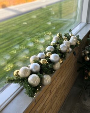 Juleguirlande pyntet med hvide, sølv og gyldne julekugler samt små LED-lys. Opsat udendørs på vindueskant og skaber elegant vinterstemning.