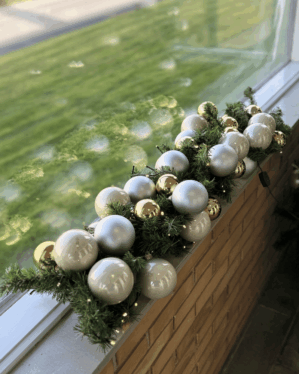 Juleguirlande pyntet med hvide, sølv og gyldne julekugler samt små LED-lys. Opsat udendørs på vindueskant og skaber elegant vinterstemning.