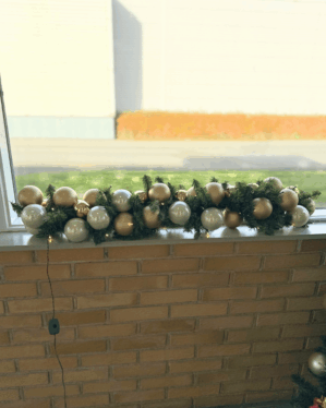 Udendørs juleguirlande pyntet med hvide og gyldne julekugler samt små LED-lys. Placeret på murstensvæg og skaber elegant, klassisk vinterudsmykning.