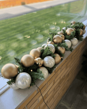 Juleguirlande pyntet med hvide og gyldne julekugler samt varme LED-lys. Placeret udendørs på vindueskant og giver et elegant, harmonisk udtryk.