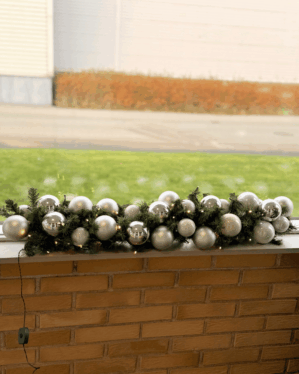 Indendørs juleguirlande pyntet med sølvfarvede og spejlblanke julekugler samt små LED-lys. Placeret på murstensvæg og skaber elegant, moderne vinterudsmykning.