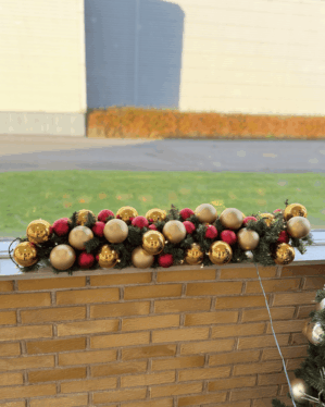 Festlig granguirlande med røde, guld og champagnefarvede julekugler på murstensgelænder i dagslys. Naturtro gran med LED-lys skaber varm julestemning udendørs.