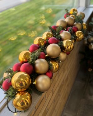 Smuk juleguirlande med røde, guld og champagnefarvede kugler på grøn gran med LED-lys. Placeret på murstensgelænder i dagslys og skaber varm julestemning.