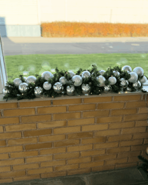 Festlig granguirlande med sølv og hvide julekugler placeret på murstensgelænder i dagslys. Naturtro gran og LED-lys skaber et elegant, vinterligt udtryk.