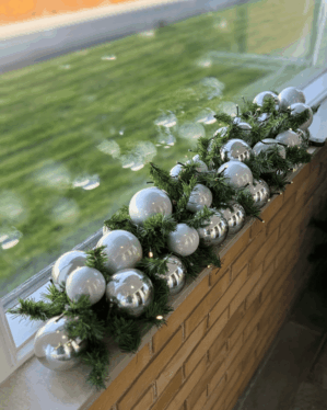 Elegant granguirlande med sølv og hvide julekugler på grønne grene. Placeret på vindueskam i dagslys, med LED-lys der skaber et vinterligt udtryk.