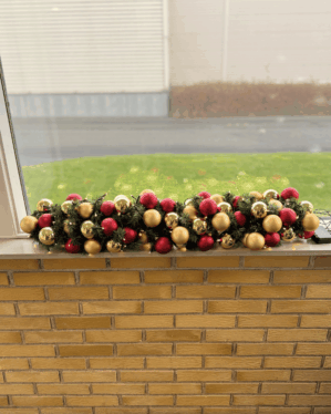 Pyntet granguirlande med røde og champagne julekugler placeret i en vindueskarm. Skaber en klassisk, varm og festlig julestemning med elegant og traditionel udstråling.