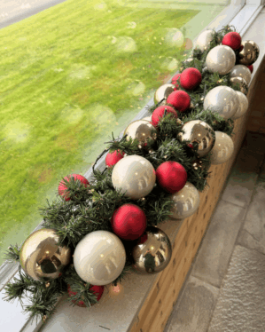 Elegant granguirlande pyntet med røde, perlemor og champagnefarvede kugler samt varm hvide LED-lys. Perfekt til vindueskarm eller reception for en moderne julestemning.