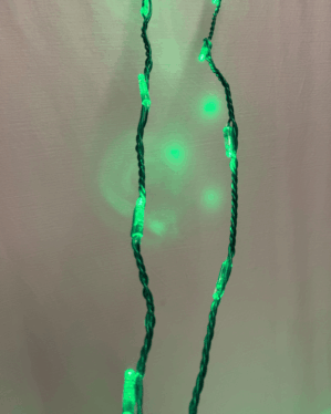 Nærbillede af grøn LED-lyskæde med tvundne mørke kabler, der hænger foran en lys baggrund og kaster bløde, grønne lysrefleksioner omkring sig.