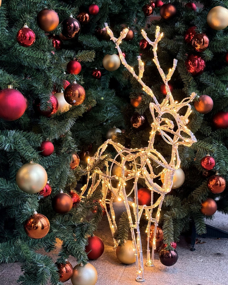 Mini stag juleudsmykning i aluminium med LED lyskæde der gemmer sig mellem nogle kunstige juletræer med julekugler.