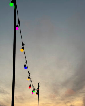 Multicolor LED party-lyskæde monteret mellem stolper udendørs, farvede pærer skaber stemningsfuld belysning mod aftenhimmel til events, haver og udeservering.