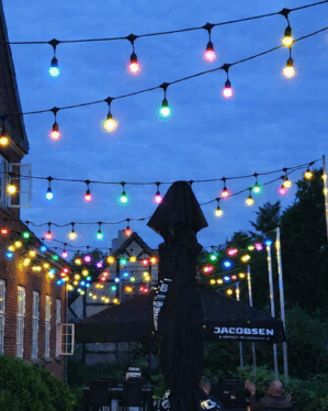 Farverige festoon lyskæder med røde, blå, grønne og gule pærer hænger over udendørs terrassemiljø ved aften og skaber stemningsfuld belysning mellem bygninger.
